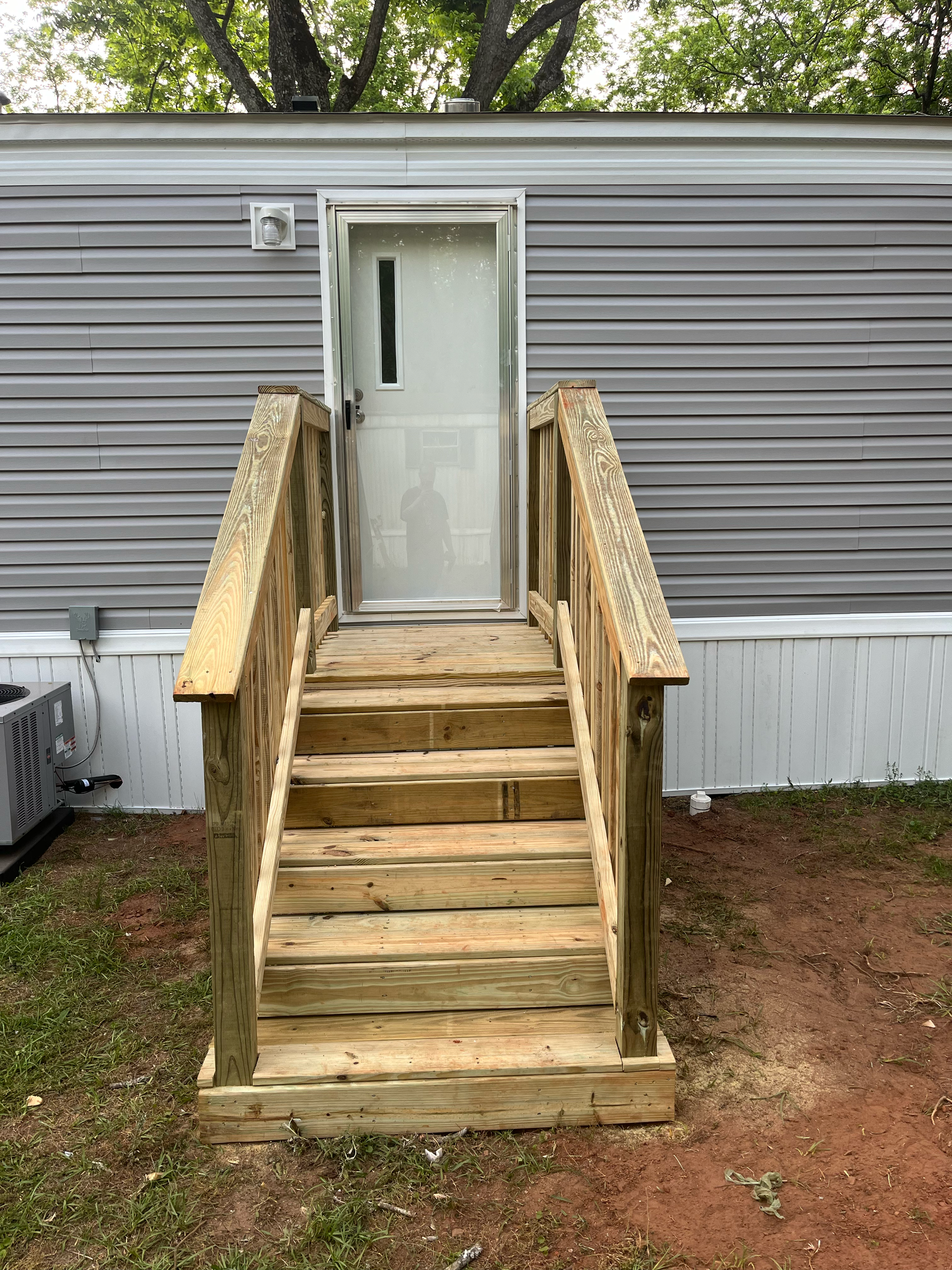 A newly built wooden staircase with railings leads up to the white door of a grey mobile home on a dirt lot.