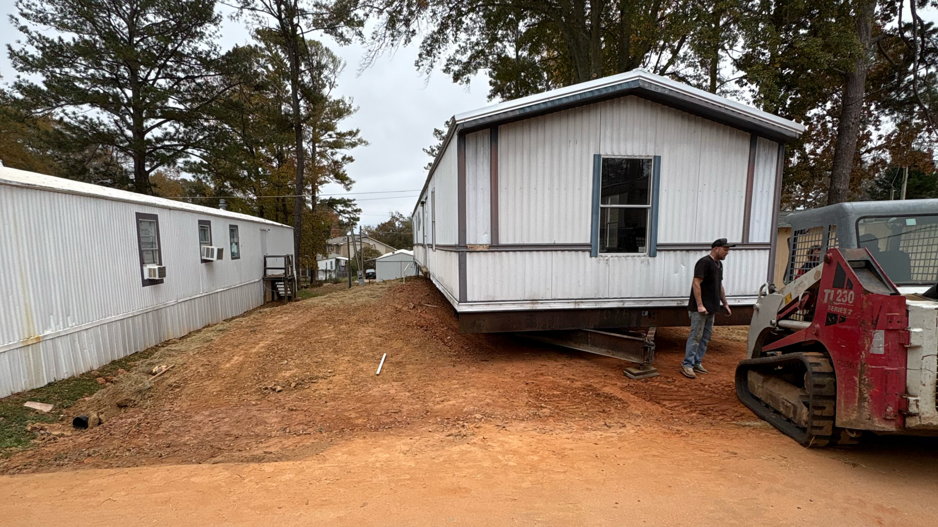Mobile home being repositioned on a dirt lot, with a worker beside a skid steer.