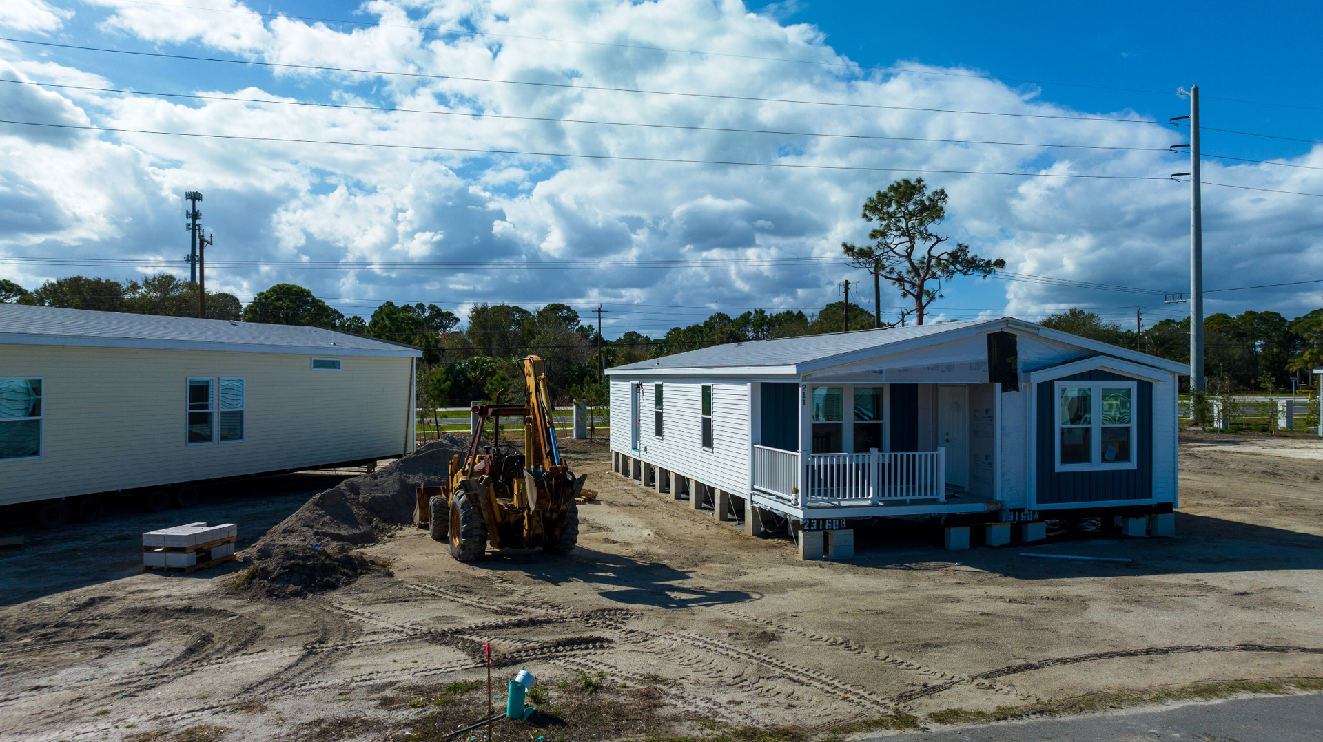 Mobile homes on a dirt lot under a cloudy sky, with a yellow excavator in the center.