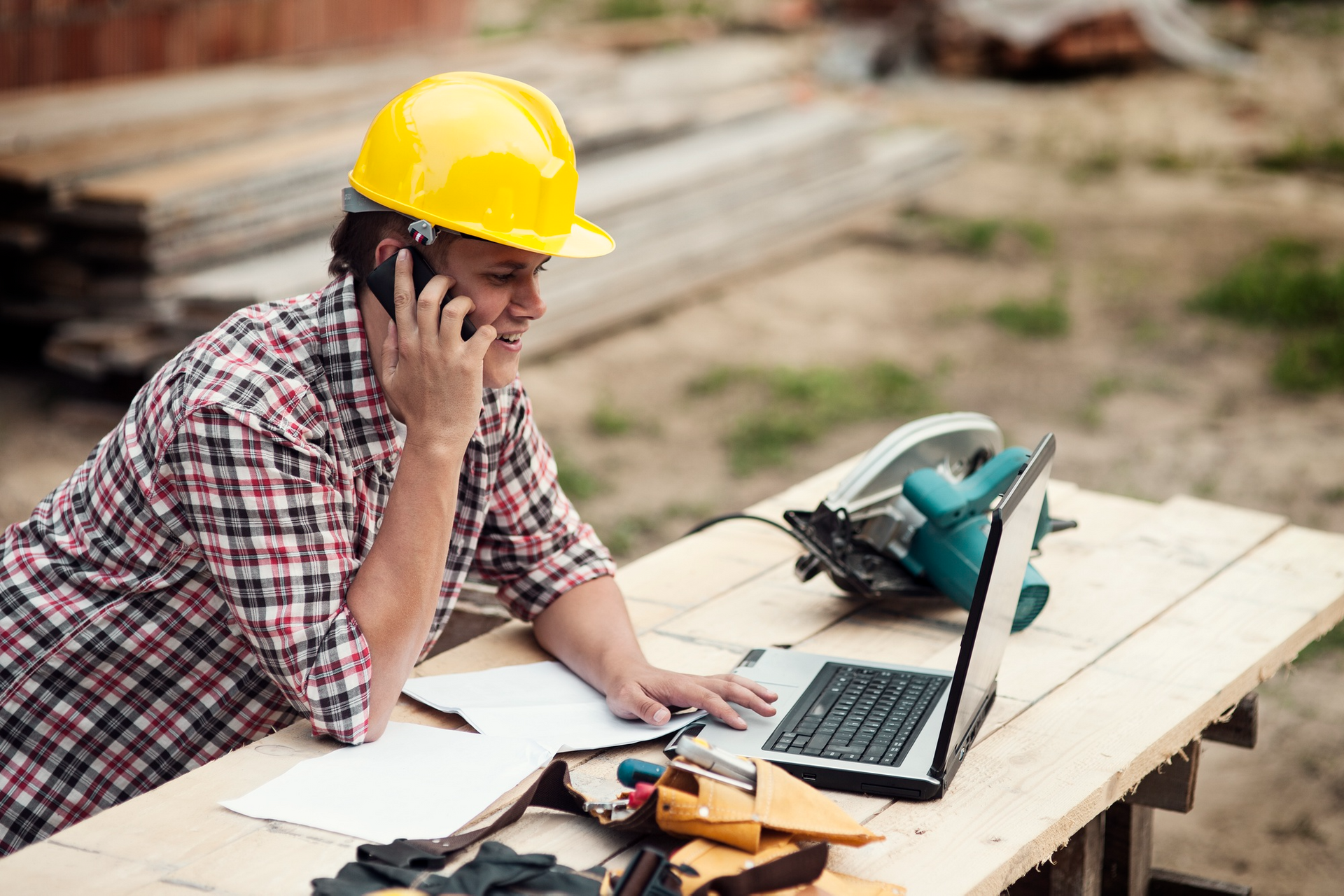 “Construction contractor using a laptop and phone at a job site to manage project plans and communic