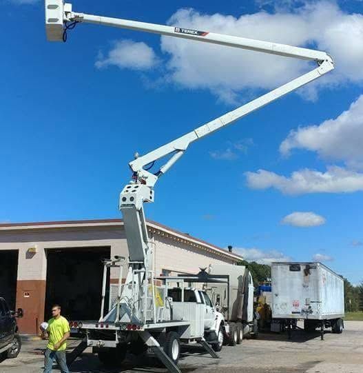 Bucket truck boom extended over a building, with workers and utility vehicles on a sunny day.