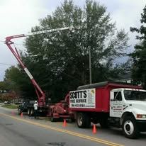 A red Scott's Tree Service truck with an extended lift boom trimming a tree on a street.