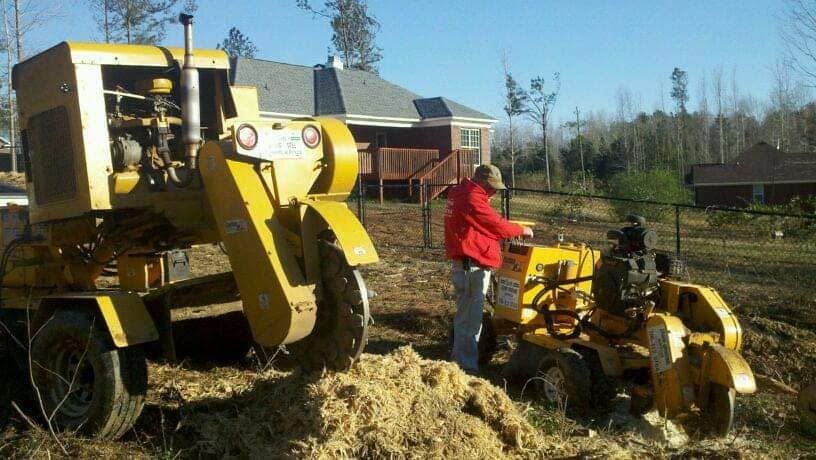 Two people standing by yellow farm machinery in a field, with a house and trees in the background.