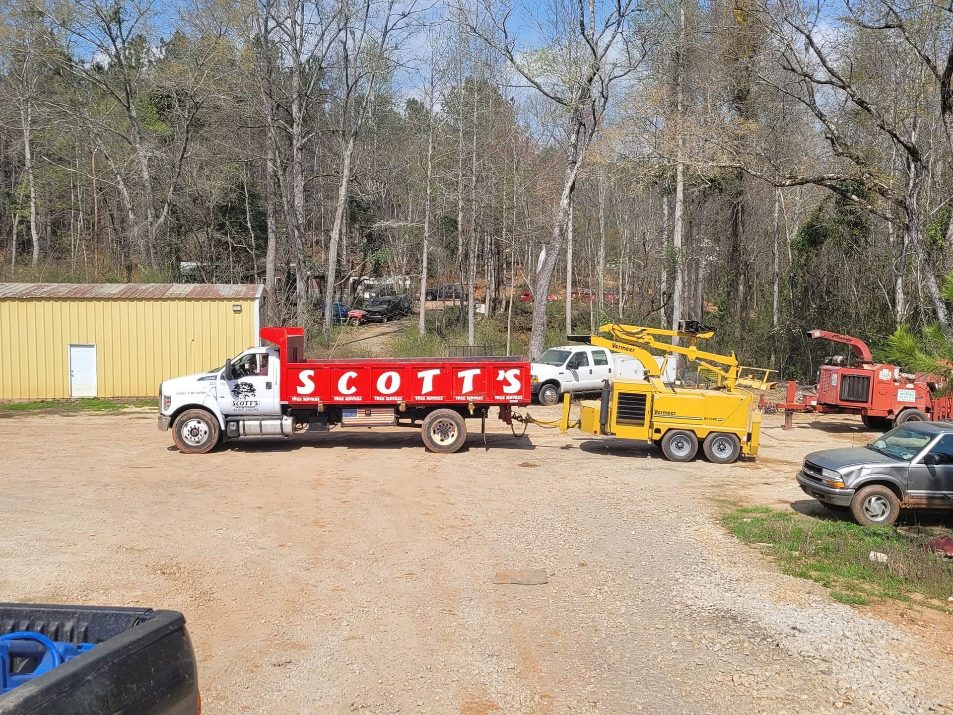 Scotts utility truck and yellow equipment parked on a gravel lot by trees and a shed