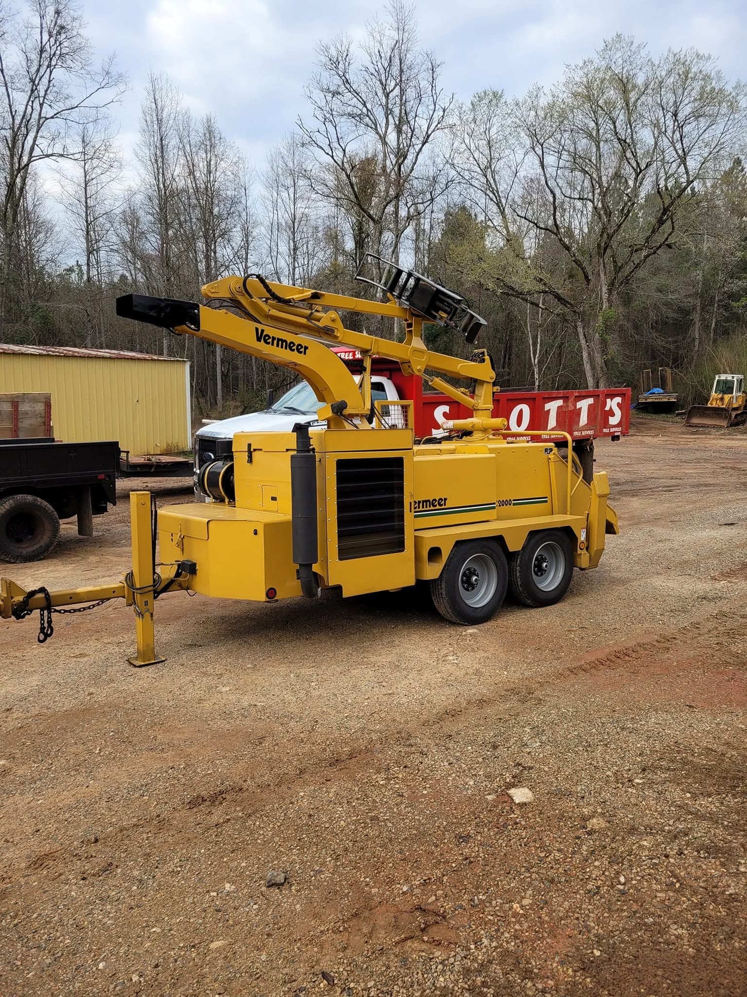Yellow trailer-mounted bucket lift parked on gravel beside a truck and red barrier, with trees in the background