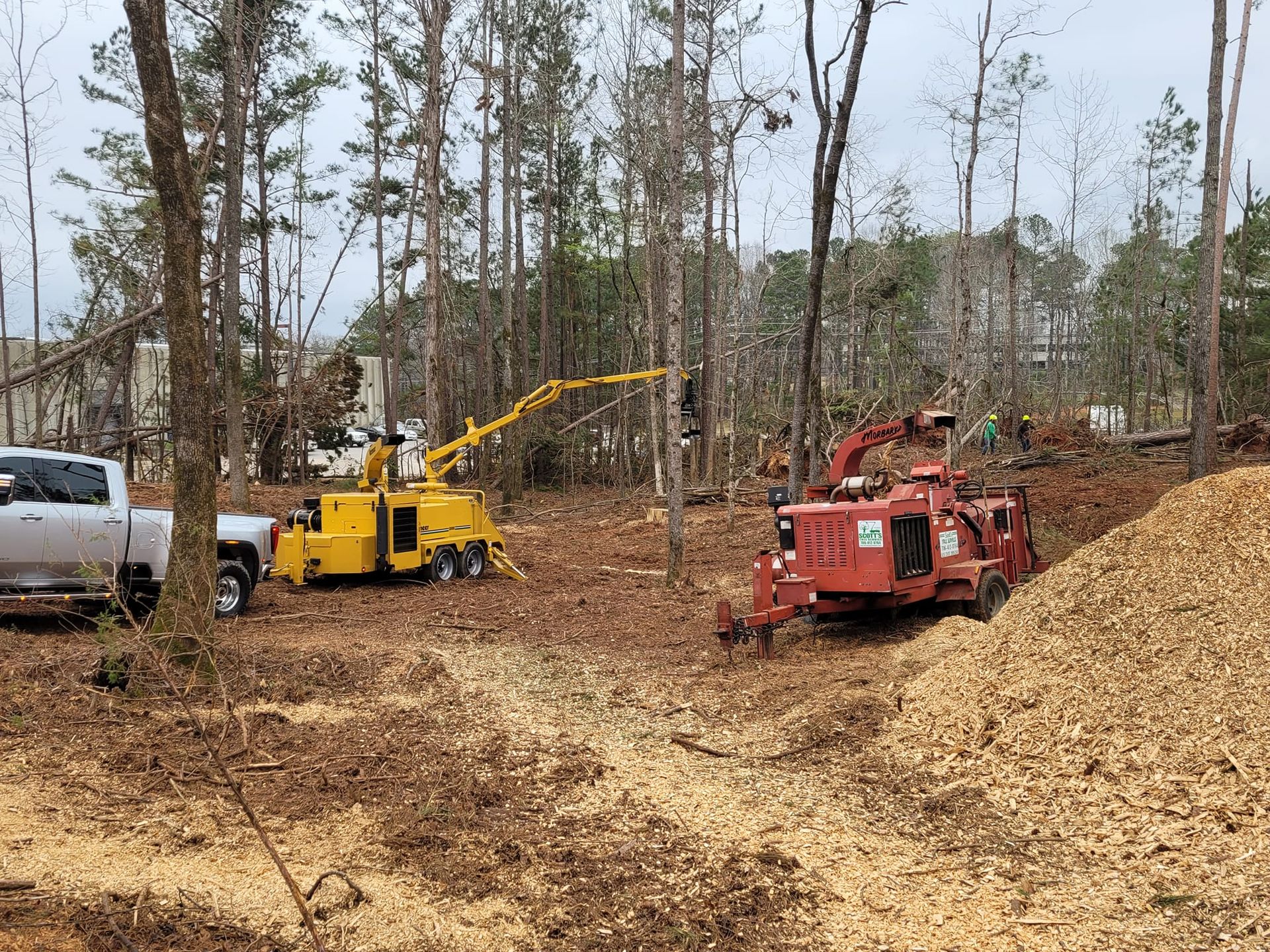 Two wood chippers in a wooded area, creating wood chips. A truck is parked nearby.