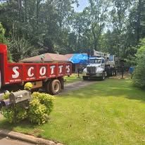 A large truck from Scotts sits near a house with other trucks and equipment, on a grassy lawn.