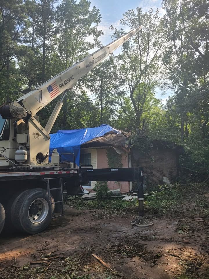 Crane truck lifting debris beside a house covered with a blue tarp in a wooded yard