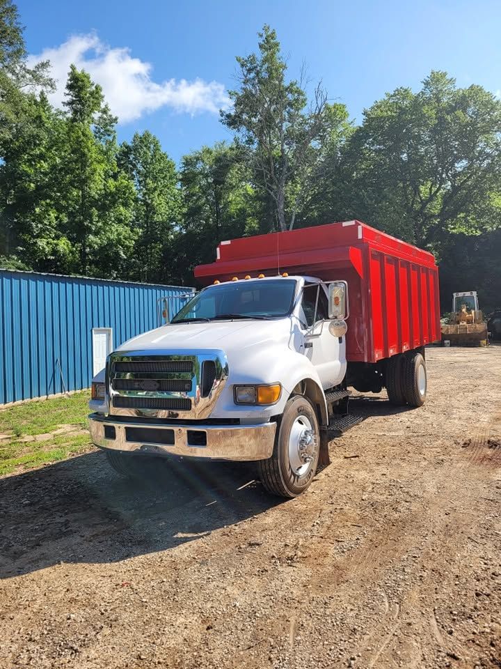 White dump truck with a red bed parked on a dirt lot beside blue fencing and trees