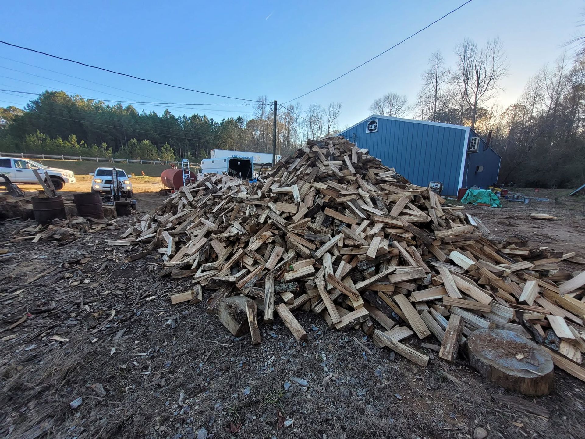 Woodpile in a muddy yard with a blue shipping container and parked vehicles in the background