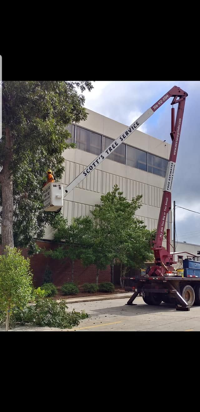 Crane lifting a white building panel beside a brick building and trees under a blue sky