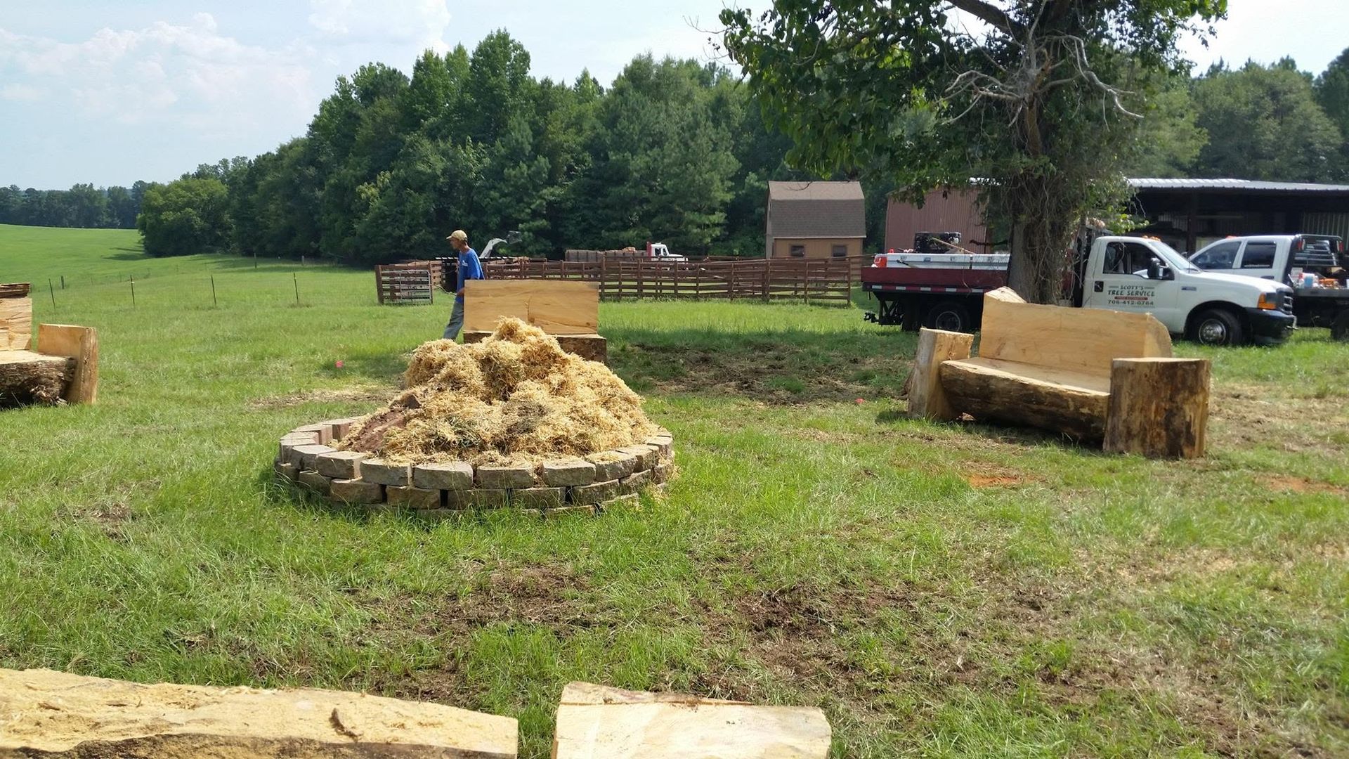 Hay bale in a grassy field with tractors, trucks, and a person nearby under a tree
