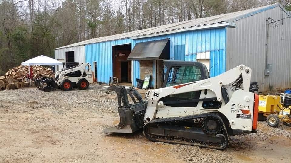 White skid steer parked outside a blue-and-gray workshop with an open bay and wood piles nearby
