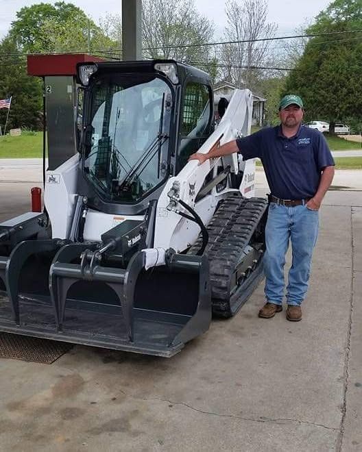 Man standing beside a white skid steer loader with black tracks on a driveway.