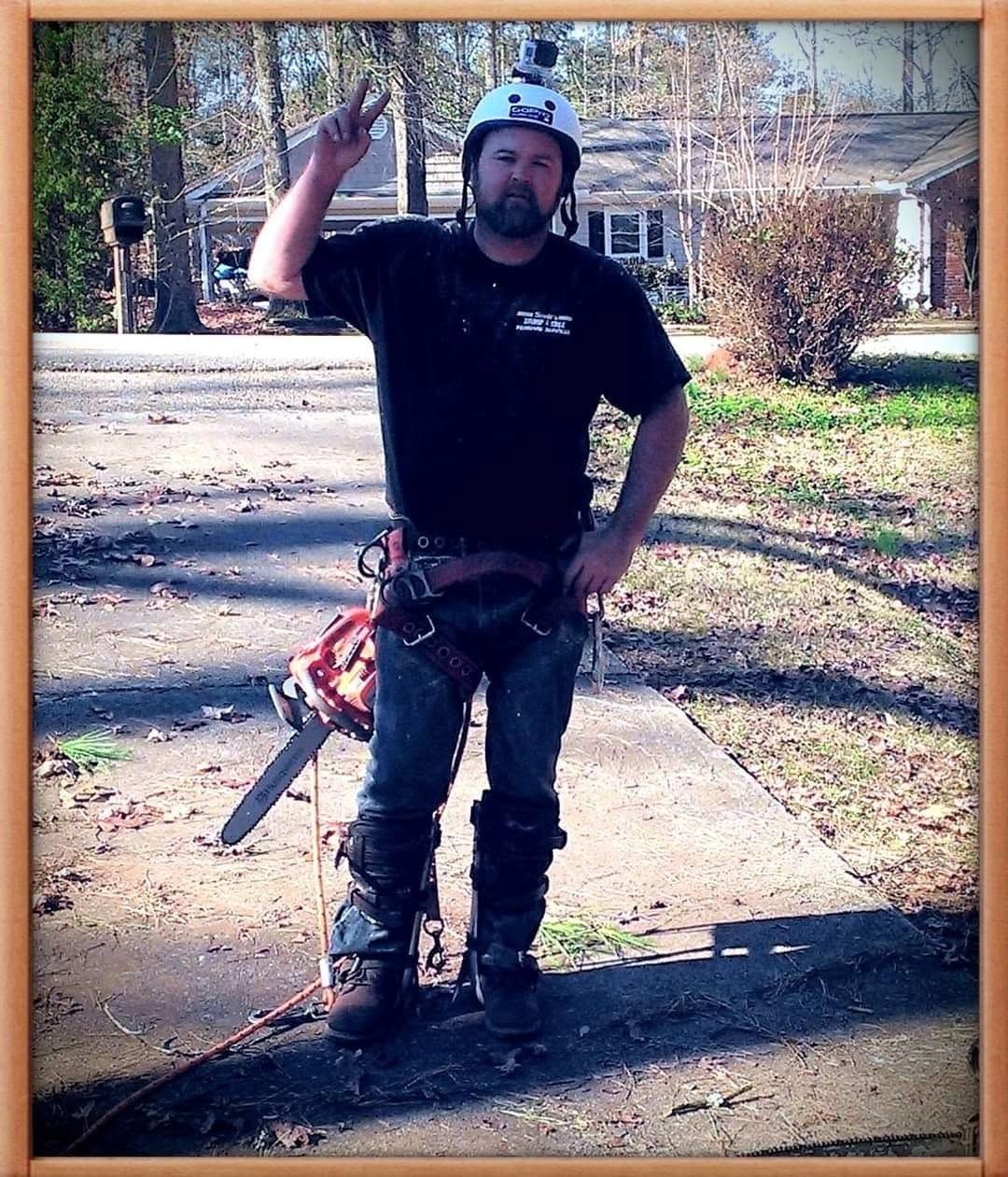 Person in a helmet stands on a driveway holding up one hand, with a chainsaw at their side.