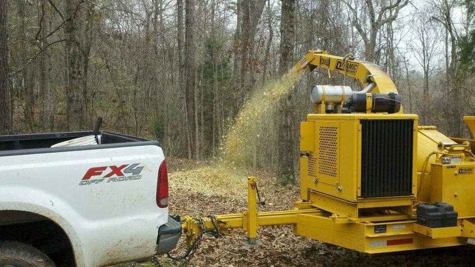 White pickup truck beside yellow wood chipper blowing wood chips in a wooded area