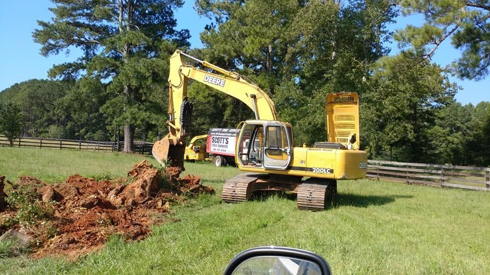 Yellow excavator digging a dirt pile in a grassy field beside a wooden fence and trees