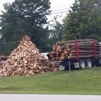 Pile of split firewood next to a truck loaded with logs, parked outdoors on a grassy verge.