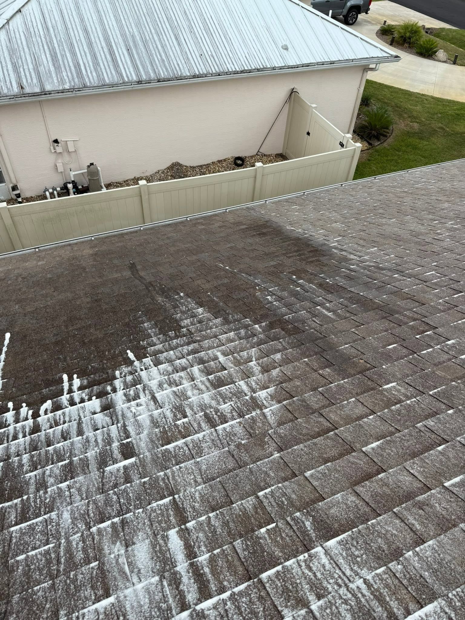 Roof shingles with white streaks or residue in the foreground, and a beige house with a metal roof in the background