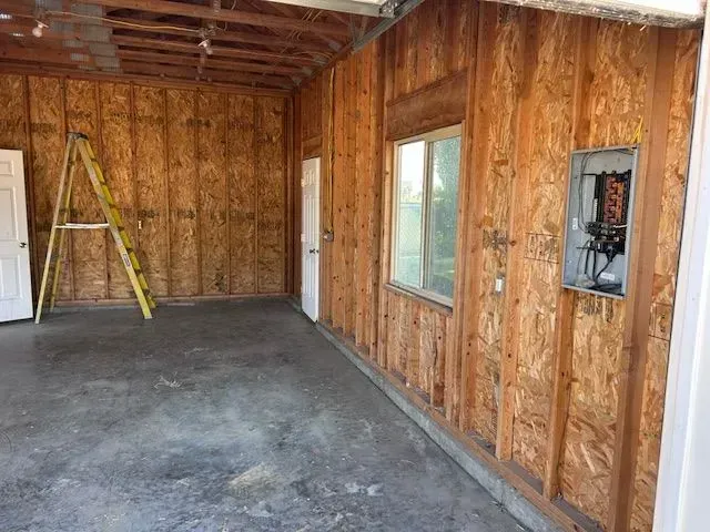 An unfinished garage interior with exposed wooden studs, a ladder, a window, and an exposed electrical breaker panel.