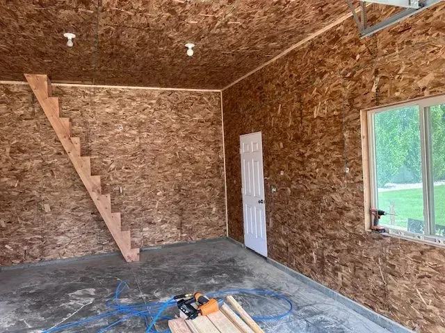 Interior of a garage with OSB-paneled walls and ceiling, a wooden staircase, a white door, and a window.