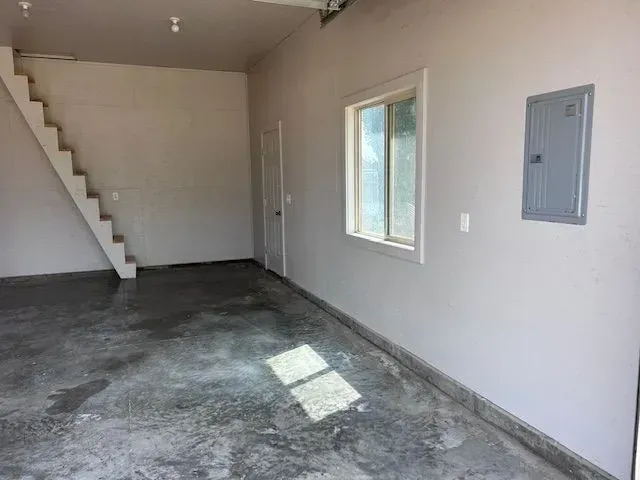 A modern, unfinished garage interior with concrete flooring, a staircase leading upward, a window, and an electrical panel.