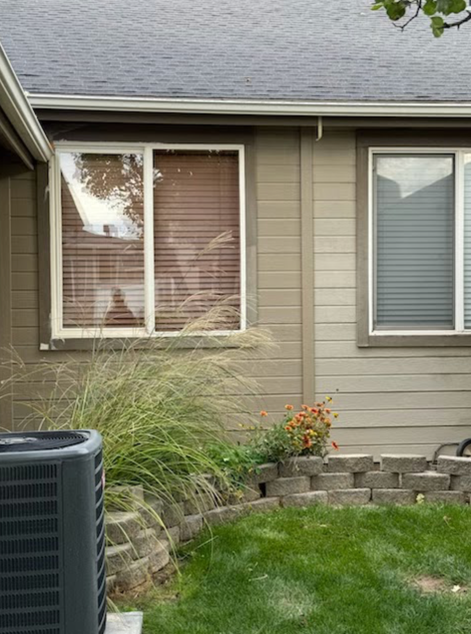 Exterior view of a tan-sided home with two windows, a partial view of an HVAC unit, and a small curved stone garden bed.