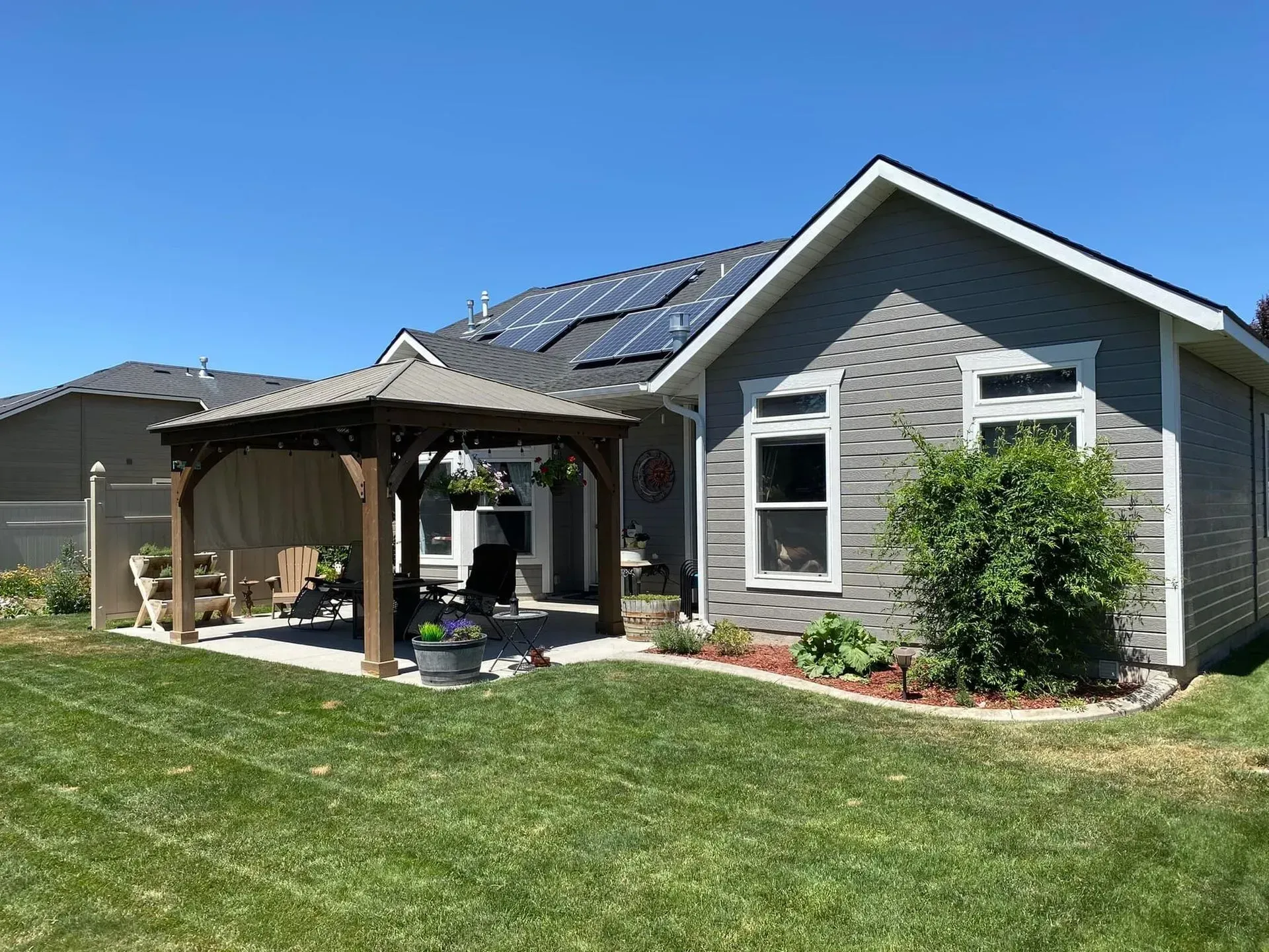A gray house with solar panels and a wooden gazebo on a concrete patio in a grassy backyard under a clear blue sky.