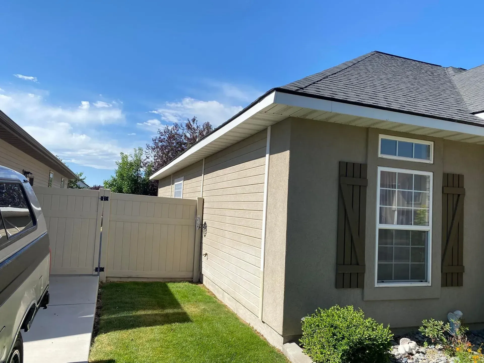 A tan house with dark brown shutters, a shingled roof, and a beige privacy fence under a clear blue sky.