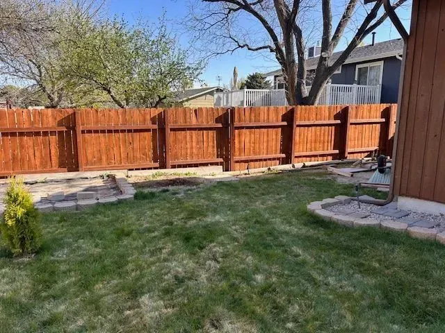 A backyard with a brown wooden fence, a patch of green grass, and small stone-bordered garden beds.