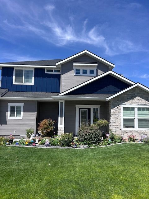 A two-story house with gray and blue siding, stone accents, a dark roof, and a front yard against a blue sky.