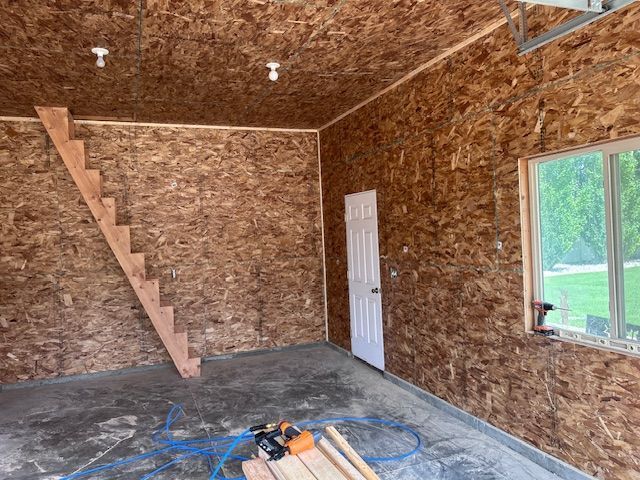 Interior of a garage with OSB-paneled walls and ceiling, a staircase against the back wall, a side door, and a window.