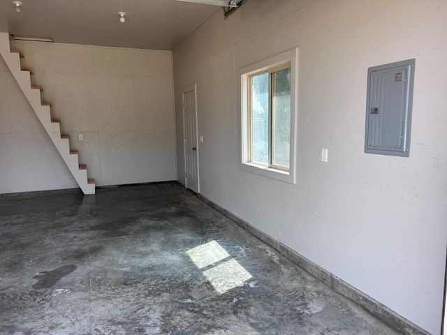 An empty garage with a concrete floor, stairs leading upward, a window on the wall, and an electrical panel.