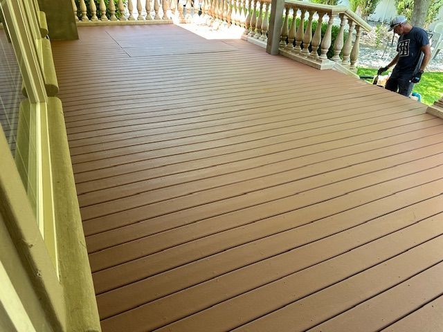 A person cleans a wide, light brown composite deck with a balustrade railing on a sunny day.