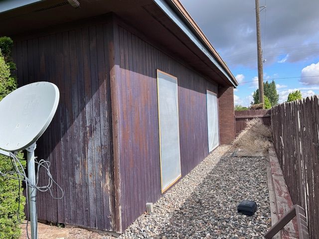 Exterior of a house side wall with dark wood siding, two rectangular panels, a gravel path, and a satellite dish.