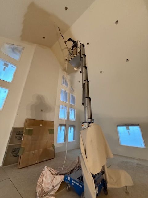 A worker in a high-reach lift paints a high ceiling in a room with multiple windows.