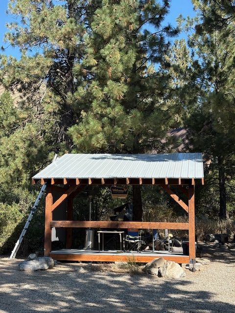 A wooden, open-air pavilion with a metal roof, furnished with a small table and chairs in a wooded, gravel-covered area.