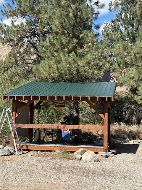 A green-roofed wooden pavilion with a small ladder standing beside it, set against a backdrop of pine trees.
