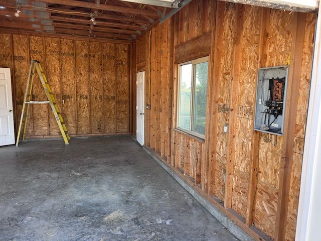 A garage interior with OSB wall panels, a yellow stepladder, a window, and an exposed electrical breaker panel.