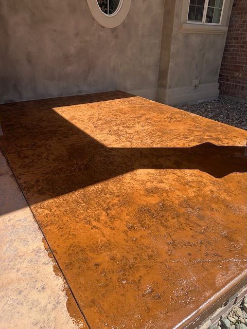 A section of concrete patio featuring a textured, rust-colored stain with a rectangular shadow cast across the surface.