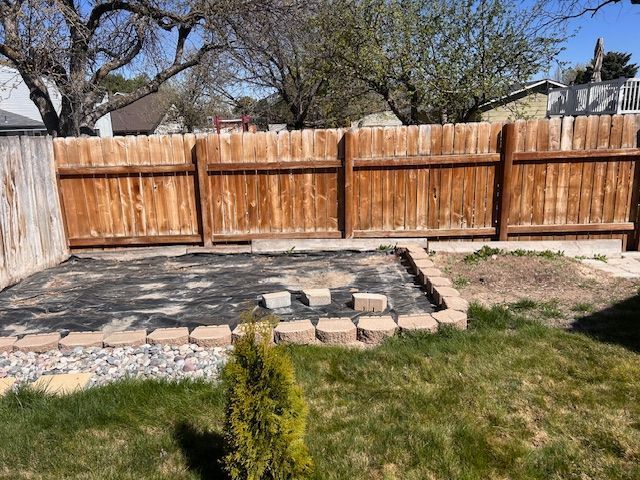 A backyard garden bed with a black landscape fabric liner, surrounded by tan concrete pavers in front of a wooden fence.