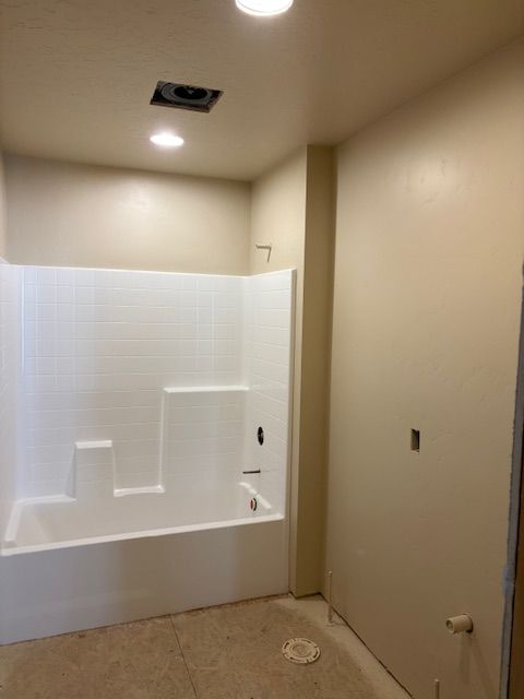 A white bathtub and shower surround in a bathroom with beige walls, recessed lighting, and an unfinished floor.