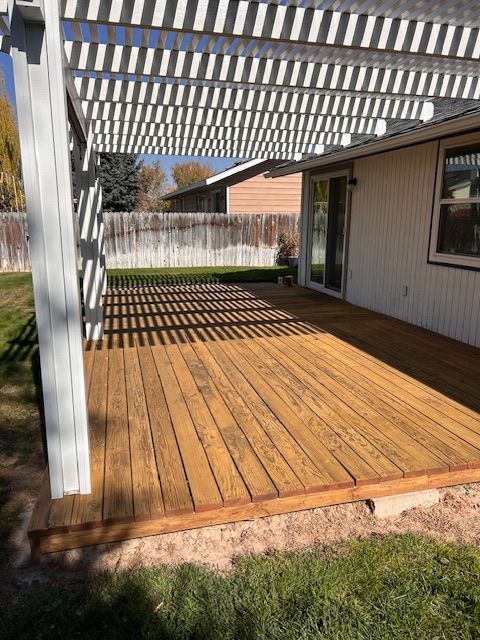 A wood deck with a pergola attached to the back of a house, overlooking a grassy yard with a wooden fence.