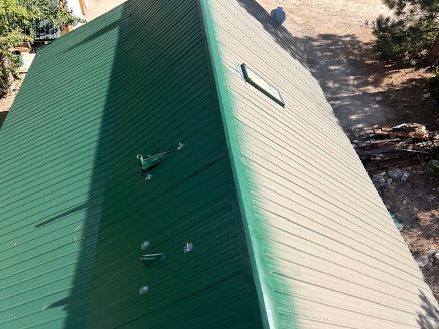 An aerial view of a pitched roof with a green metal panel side and a tan shingled side, featuring a small skylight.
