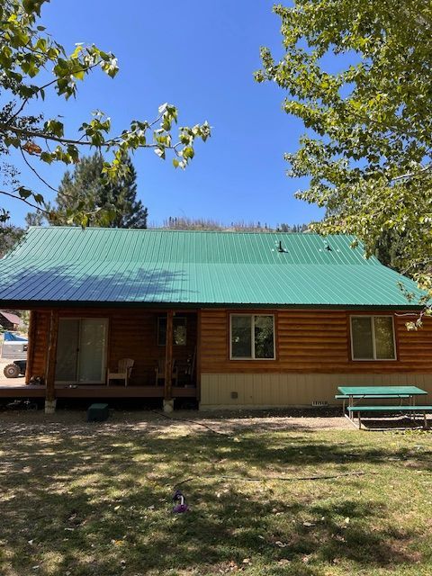 A log cabin with a green metal roof, a front porch, and a picnic table on the lawn under a clear blue sky.