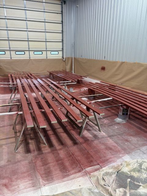 Long wooden planks painted in a reddish-brown hue resting on metal sawhorses inside a large warehouse workshop.