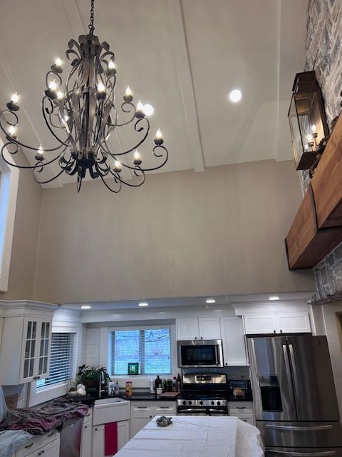 A kitchen with white cabinets, a large farmhouse sink, a stainless steel refrigerator, and a chandelier under high ceilings.