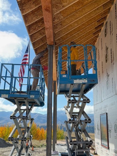 Two construction workers on scissor lifts work on the wooden rafters of a building frame near an American flag.