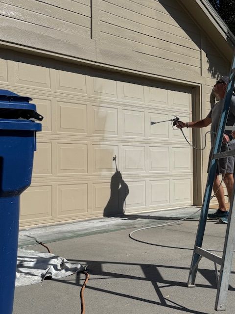 A person on a ladder uses a spray paint gun to paint a beige garage door.