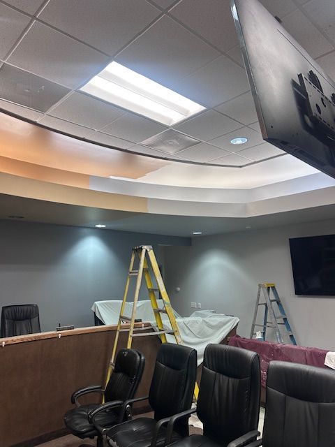 An office space under renovation with ladders, black office chairs, a drop cloth, and partially painted ceiling details.